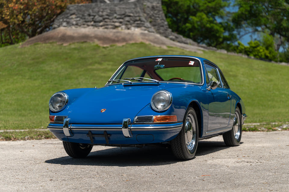 Gulf Blue, glowing blue 1966 Porsche 911 in front of a hill with stone foundation above. Porsche Leasing with Premier. This Porsche alnost looks like it is smiling.