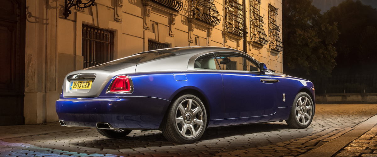 Right rear view of Rolls-Royce Wraith, night.Blue and silver two door. City Street Scene, wrought iron windor grills italian style. Rolls-Royce Leasing